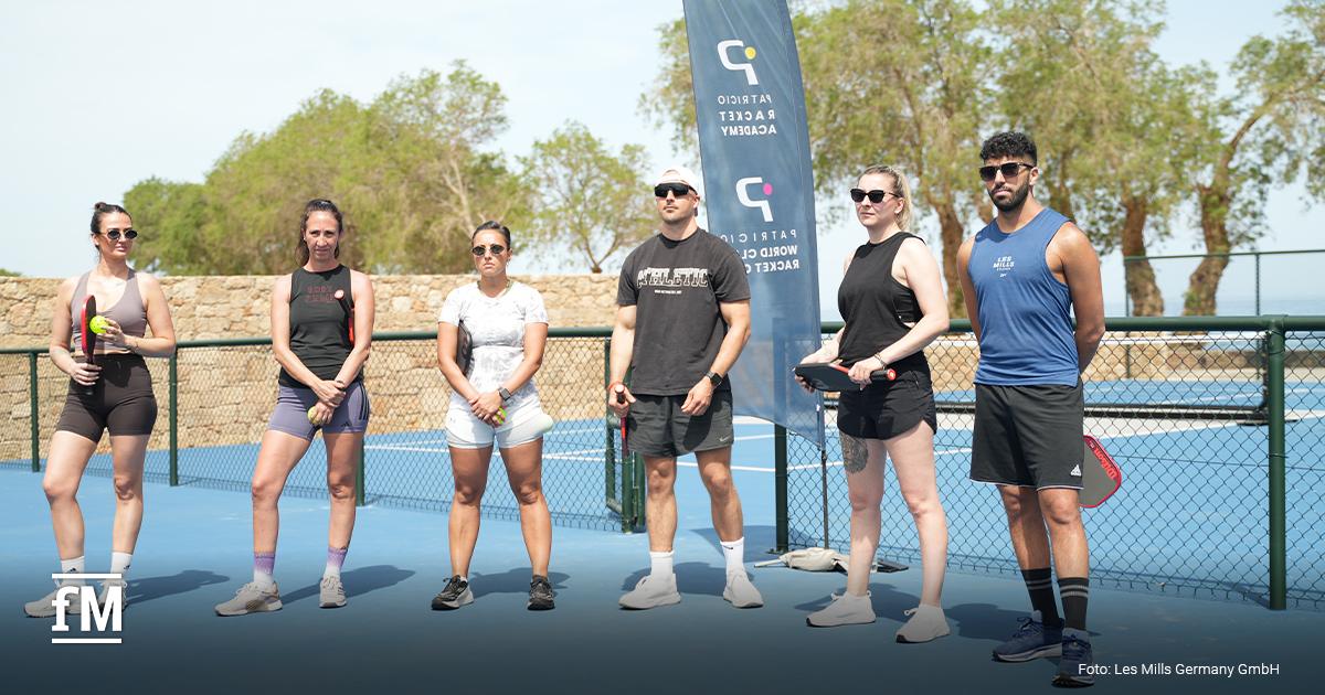 Sechs Personen stehen auf einem blauen Sportplatz und halten Pickleballschläger. Im Hintergrund steht eine blaue Fahne mit der Aufschrift „Patricio Racket Academy“ und „Patricio World Class Racket Club“. Unten links ist das fM-Logo zu sehen. Unten rechts steht „Foto: Les Mills Germany GmbH“.