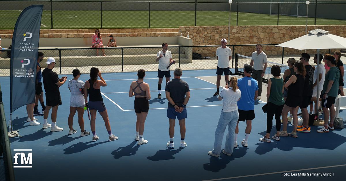 Eine Gruppe steht auf einem blauen Court im Kreis um drei Trainer. Links steht eine blaue Fahne mit der Aufschrift „Patricio Racket Academy“ und „Patricio World Class Racket Club“. Unten links ist das fM-Logo zu sehen. Unten rechts steht „Foto: Les Mills Germany GmbH“.