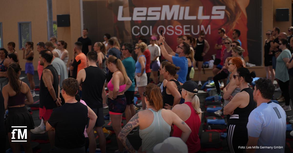 Teilnehmende stehen im Freien vor einer großen Wand mit der Aufschrift „Les Mills Colosseum“. Unten links ist das fM-Logo zu sehen. Unten rechts steht „Foto: Les Mills Germany GmbH“.