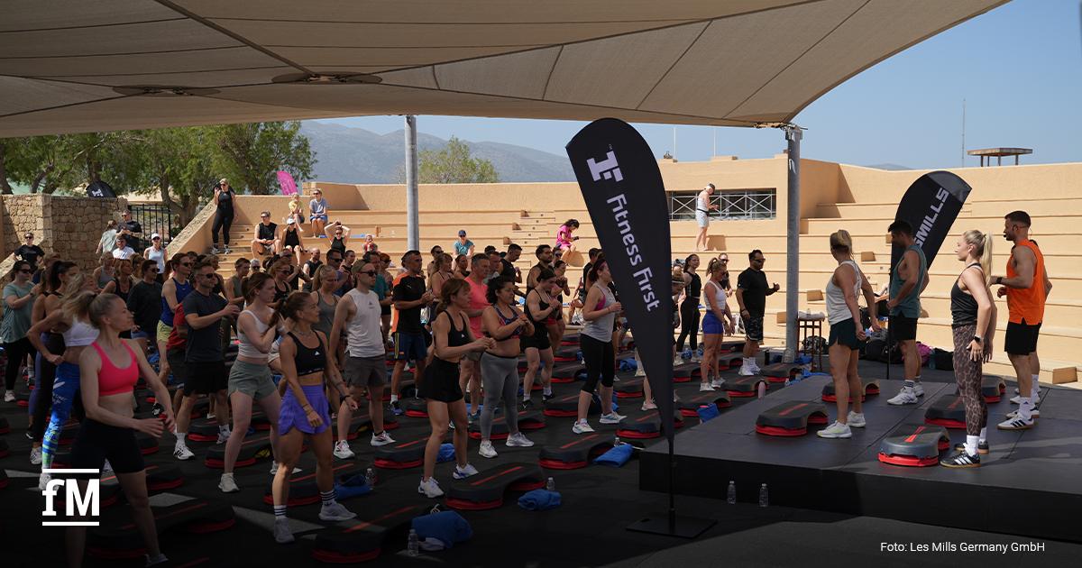 Eine Gruppe trainiert im Freien auf Stepboards vor einer Bühne. Zwei schwarze Fahnen mit den Aufschriften „Fitness First“ und „Les Mills“ stehen neben der Bühne. Unten links ist das fM-Logo zu sehen. Unten rechts steht „Foto: Les Mills Germany GmbH“.