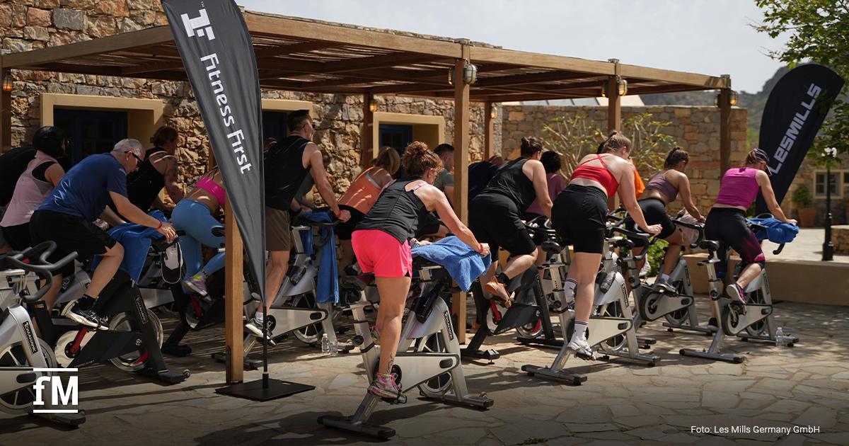 Mehrere Personen trainieren auf Indoorbikes unter einer Holzüberdachung im Außenbereich. Links steht eine schwarze Fahne mit der Aufschrift „Fitness First“, rechts eine schwarze Fahne mit der Aufschrift „Les Mills“. Unten links ist das fM-Logo zu sehen. Unten rechts steht „Foto: Les Mills Germany GmbH“.