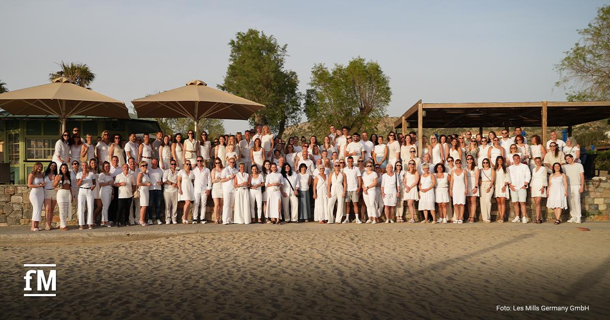 Eine große Gruppe steht am Strand vor Bäumen, Sonnenschirmen und Gebäuden. Viele Personen tragen weiße Kleidung. Unten links ist das fM-Logo zu sehen. Unten rechts steht „Foto: Les Mills Germany GmbH“.