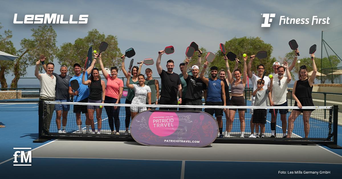 les-mills-week-kreta-titel Eine Gruppe steht auf einem blauen Court hinter einem Netz und hält Pickleballschläger und Bälle hoch. Oben links steht das Logo „Les Mills“. Am Netz hängt ein Banner mit der Aufschrift „Patricio Travel“ und „patriciotravel.com“. Unten links ist das fM-Logo zu sehen. Unten rechts steht „Foto: Les Mills Germany GmbH“.