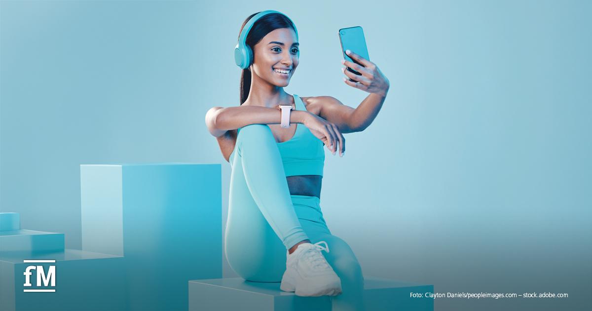 MANAGEMENT Impuls Influencer Eine Frau in heller Sportbekleidung sitzt auf einem Podest und hält ein Smartphone für ein Selfie vor sich. Sie trägt große hellblaue Kopfhörer und eine Smartwatch. Unten links ist das weiße fM-Logo zu sehen.