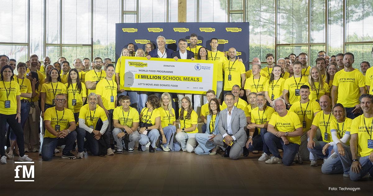 Technogym Let’s Move & Donate Food Gruppenfoto in einer Halle mit großer Fensterfront. Im Vordergrund und in der Bildmitte halten mehrere Personen einen großen symbolischen Scheck mit den Aufschriften „Technogym“, „World Food Programme“ und „1 MILLION SCHOOL MEALS“. Zahlreiche Personen tragen gelbe T-Shirts mit dem Text „LET'S MOVE FOR A BETTER WORLD“. Im Hintergrund steht eine Technogym-Bannerwand mit Logos.