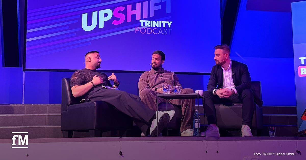 Drei Männer sitzen auf einer Bühne vor einer Leinwand mit den Schriftzügen „UPSHIFT“ und „TRINITY PODCAST“.