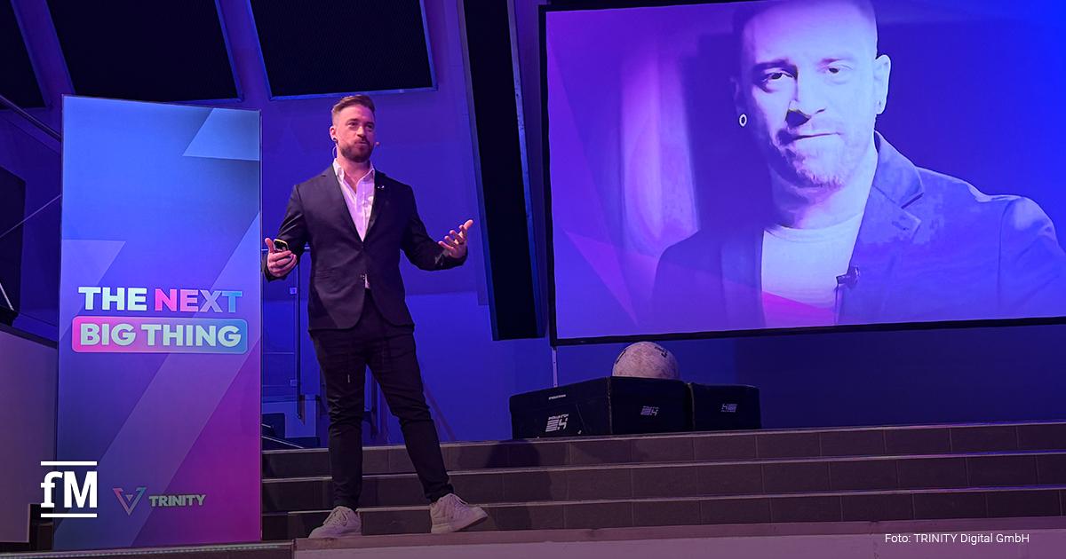 Ein Mann steht auf einer Bühne neben einem Rednerpult mit „THE NEXT BIG THING“. Hinter ihm eine große Leinwand mit einem Porträtfoto.