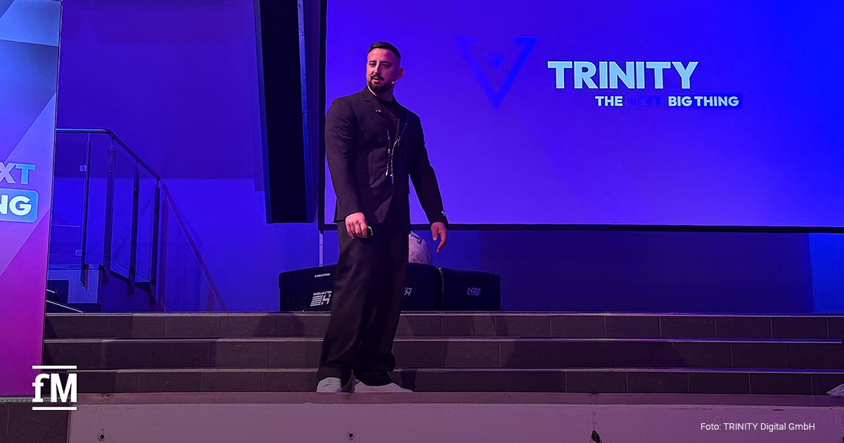 Ein Mann steht auf einer Bühne vor einer Leinwand mit den Schriftzügen „TRINITY“ und „THE NEXT BIG THING“.