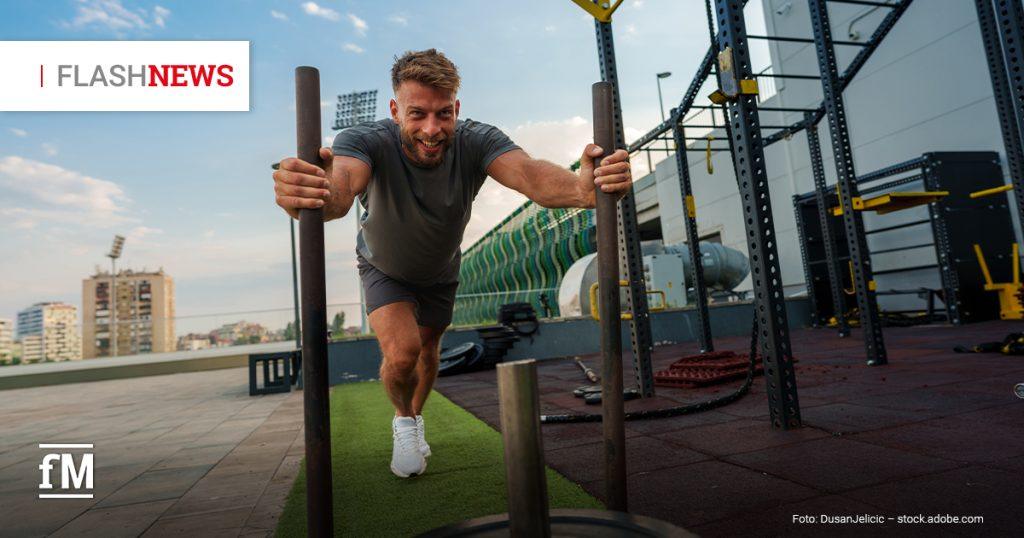 Mann schiebt im Außenbereich eines Fitnessstudios einen Gewichtsschlitten über Kunstrasen und blickt lächelnd nach vorn. Oben links steht „FLASH NEWS“, im Hintergrund sind Trainingsgeräte und Gebäude zu sehen.