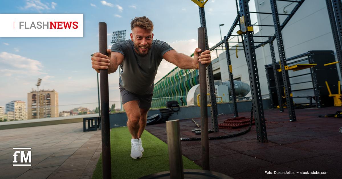 Fitness Flash News 07/2026 KW 28 Mann schiebt im Außenbereich eines Fitnessstudios einen Gewichtsschlitten über Kunstrasen und blickt lächelnd nach vorn. Oben links steht „FLASH NEWS“, im Hintergrund sind Trainingsgeräte und Gebäude zu sehen.