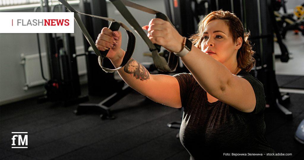 Eine Frau trainiert in einem Fitnessstudio mit Schlingentrainern. Im Hintergrund sind Krafttrainingsgeräte zu sehen.