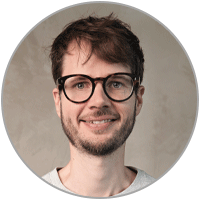 Porträt von Bernd Hoffschlag vor hellem, unscharfem Hintergrund. Er trägt eine dunkle Brille, ein helles T-Shirt und lächelt direkt in die Kamera.