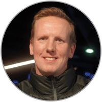 Porträt von Nils Kuprat vor dunklem Hintergrund mit unscharfen Lichtpunkten. Er trägt eine dunkle Jacke mit Reißverschluss und blickt in die Kamera.