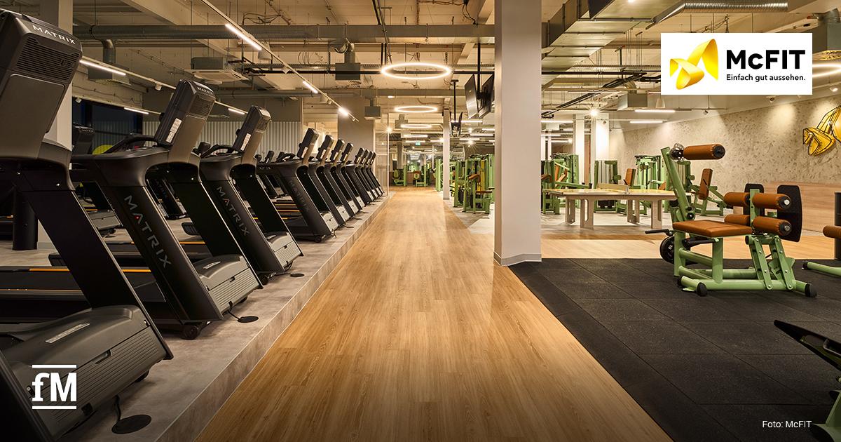 McFit Modernisierung Bochum Hofstede Reihe schwarzer Matrix-Laufbänder links im Vordergrund, mittig ein breiter Holzweg, rechts Krafttrainingsgeräte in Grün und Braun. Oben rechts McFIT-Logo mit dem Schriftzug „Einfach gut aussehen.“ Unten links fM-Logo, unten rechts der Hinweis „Foto: McFIT“.