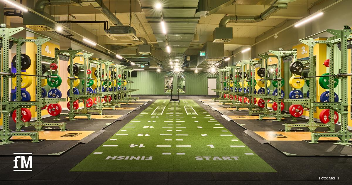 Mehrere Half Racks stehen links und rechts eines grünen Kunstrasens mit Markierungen und den Aufschriften „START“ und „FINISH“. Bunte Gewichtsscheiben hängen an den Racks.