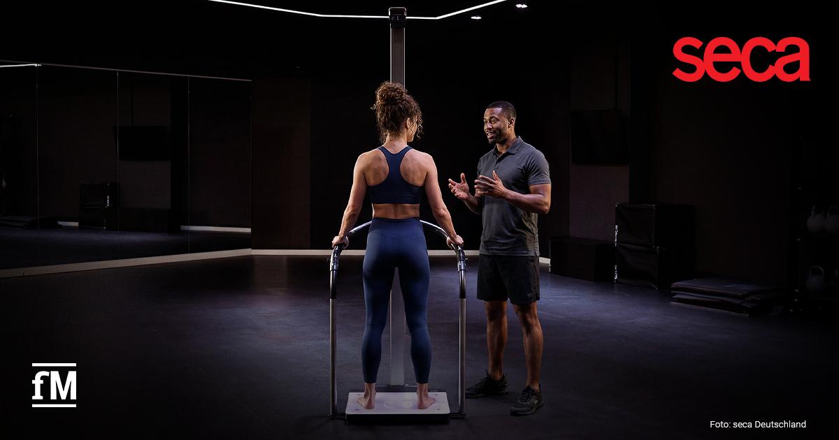 seca Advertorial Eine junge Frau in Sportkleidung steht auf einer BIA-Waage. Rechts neben ihr ein Trainer. Die Umgebung ist modern und in dunklen Farben gehalten.