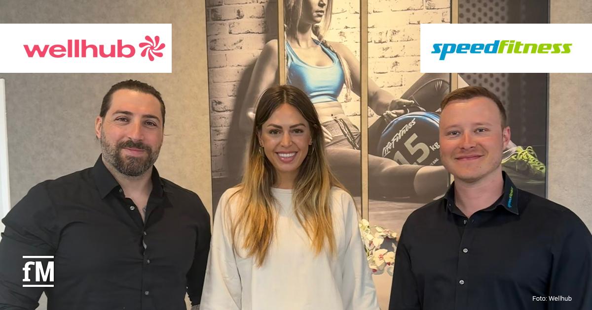 Speedfitness x Wellhub Drei lächelnde Personen stehen in einem modern eingerichteten Raum vor einem großformatigen Wandbild mit Fitnessmotiv.