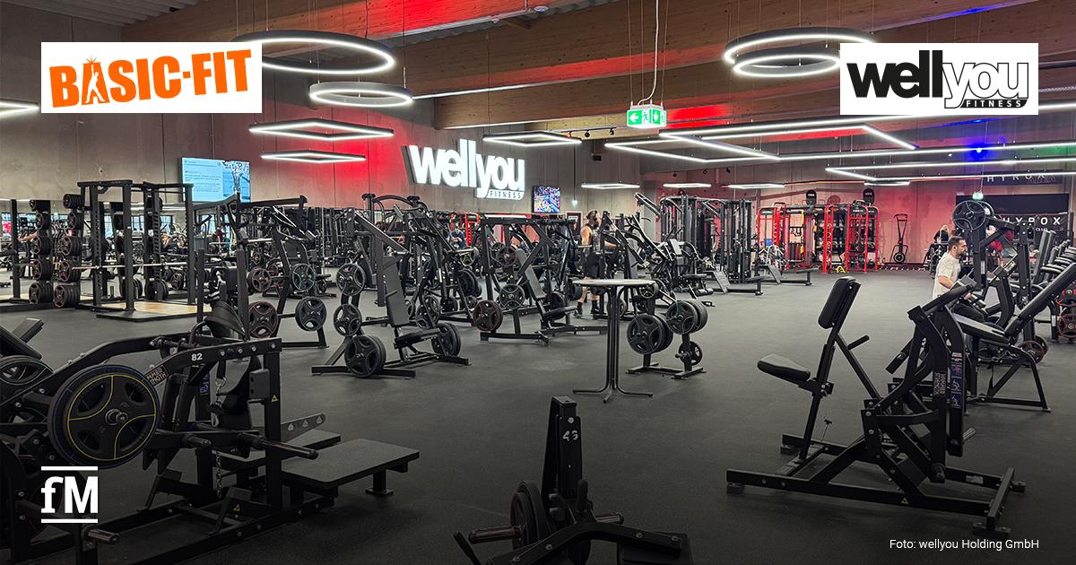 Basic-Fit kauft wellyou Großer Fitnessstudiobereich mit zahlreichen Krafttrainingsgeräten und Freihantelstationen. Im Hintergrund ist ein beleuchtetes „wellyou Fitness“-Logo an der Wand zu sehen. An der Decke hängen ringförmige und rechteckige Leuchten. Oben links befindet sich das Basic-Fit-Logo, oben rechts das wellyou-Fitness-Logo.