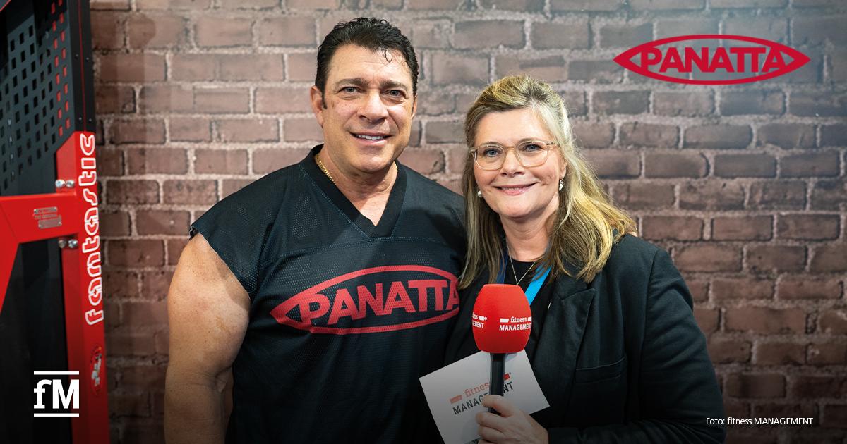 Interview Panatta Bob Cicherillo Links im Vordergrund steht ein Mann in schwarzem Oberteil mit rotem Panatta-Schriftzug neben einer Frau mit Brille und rotem Mikrofon vor einer Backsteinwand.