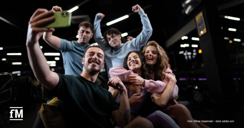Fünf junge Erwachsene sitzen gemeinsam in einem Fitnessstudio und machen ein Gruppen-Selfie.