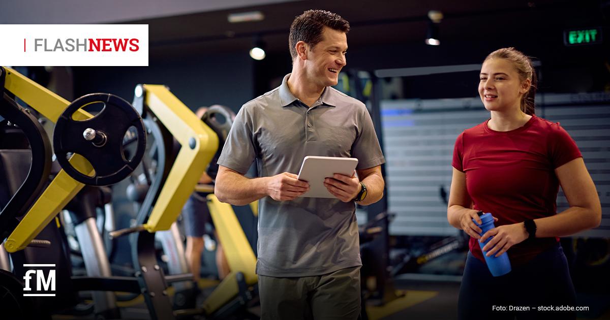 Fitness Flash News #0872026 KW 33 Ein Trainer mit Tablet spricht lächelnd mit einer Sportlerin, die eine Trinkflasche hält, zwischen Kraftgeräten in einem Fitnessstudio.