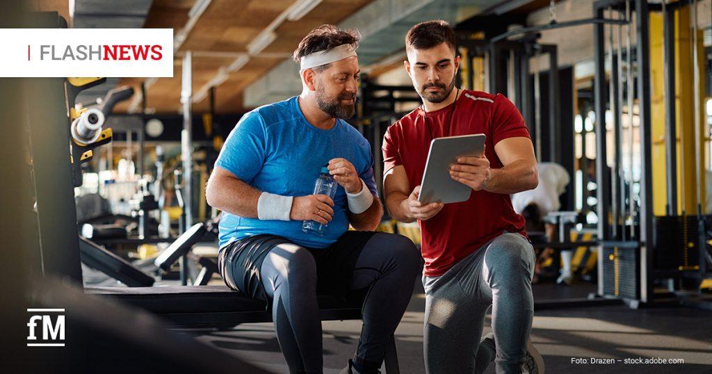 Zwei Männer in Sportkleidung sitzen in einem Fitnessstudio. Einer hält eine Wasserflasche, während der andere ihm etwas auf einem Tablet zeigt. Links oben steht „FLASHNEWS“.
