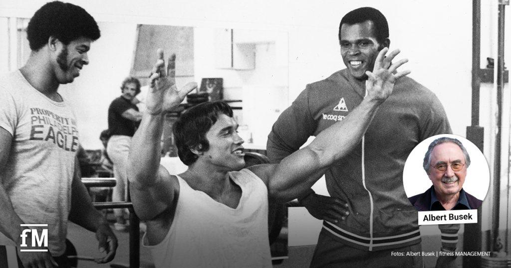 Drei Männer in einem Fitnessstudio, zwei davon lächelnd im Gespräch; rechts ein Porträt von Albert Busek.