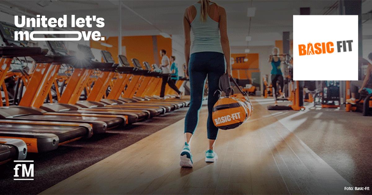 Basic-Fit unterstützt „United let’s move.“ Frau mit Basic-Fit-Sporttasche geht zwischen Laufbändern durch ein Fitnessstudio