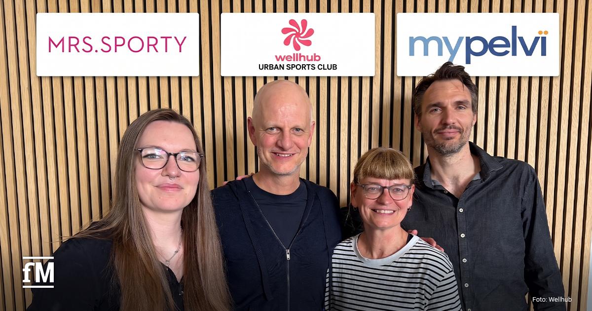 Wellhub Urban Sports Club stärkt Frauengesundheit mit Mrs.Sporty und MyPelvi Vier Personen stehen vor einer Holzwand unter den Logos von Mrs.Sporty, Wellhub Urban Sports Club und MyPelvi.
