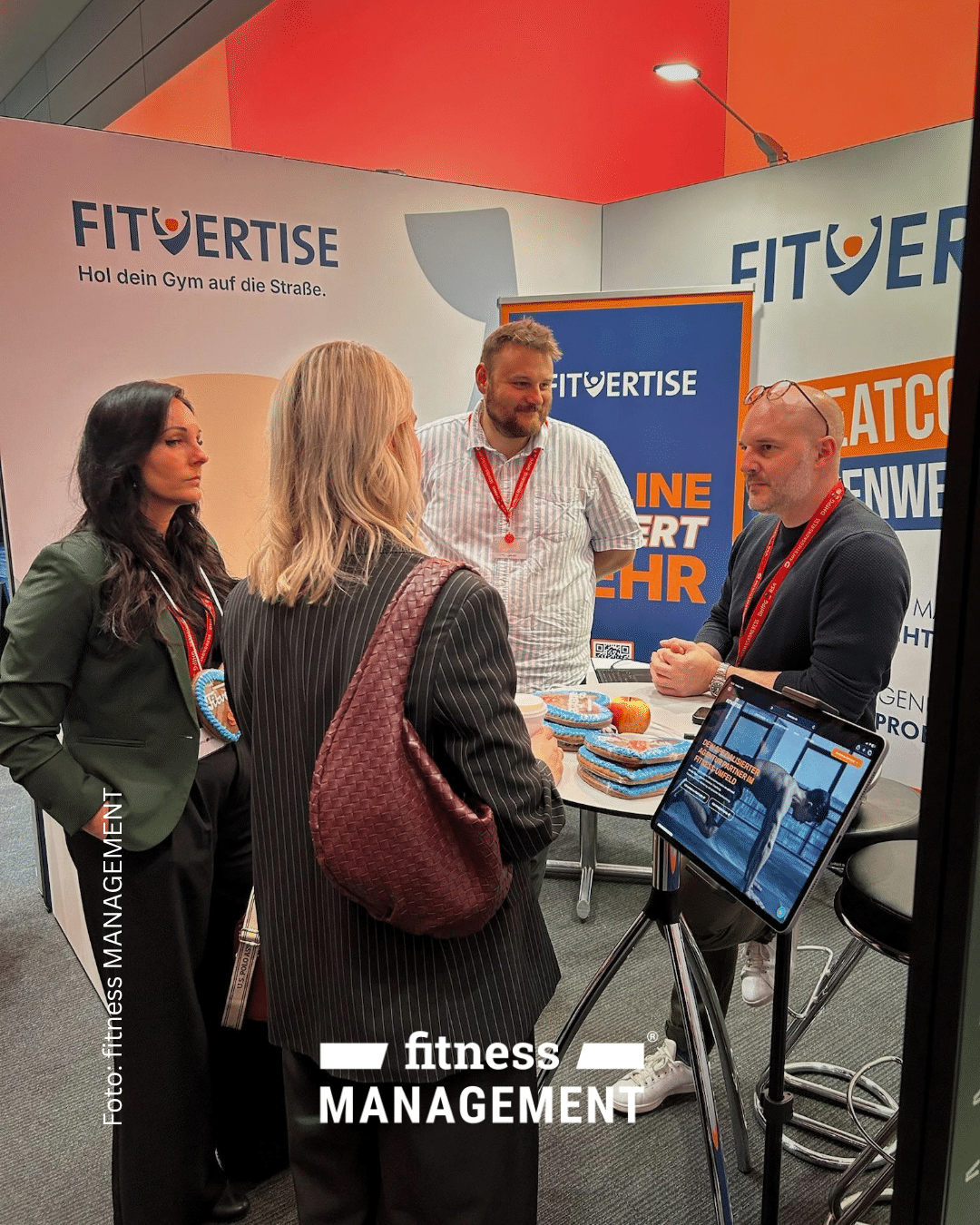 Vier Personen unterhalten sich am Messestand von FITVERTISE. Neben einem Stehtisch zeigt ein Bildschirm digitale Marketinginhalte.