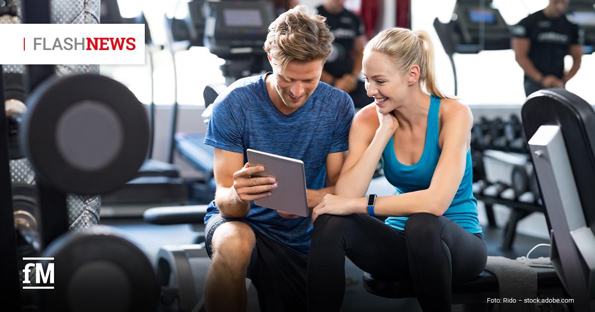 Fitness Flash News #10/2026 KW42 Ein lächelnder Mann und eine Frau in Sportkleidung sitzen nebeneinander im Fitnessstudio und schauen gemeinsam auf ein Tablet. Im Hintergrund sind Trainingsgeräte zu sehen. Oben links steht „FLASHNEWS“.