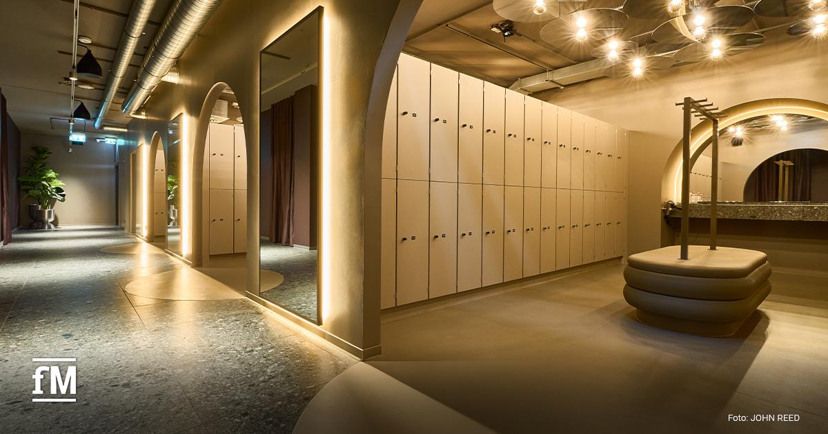Eleganter Umkleidebereich mit beigen Schließfächern, beleuchteten Ganzkörperspiegeln, Rundbögen und gepolsterter Sitzbank in warmem Licht.