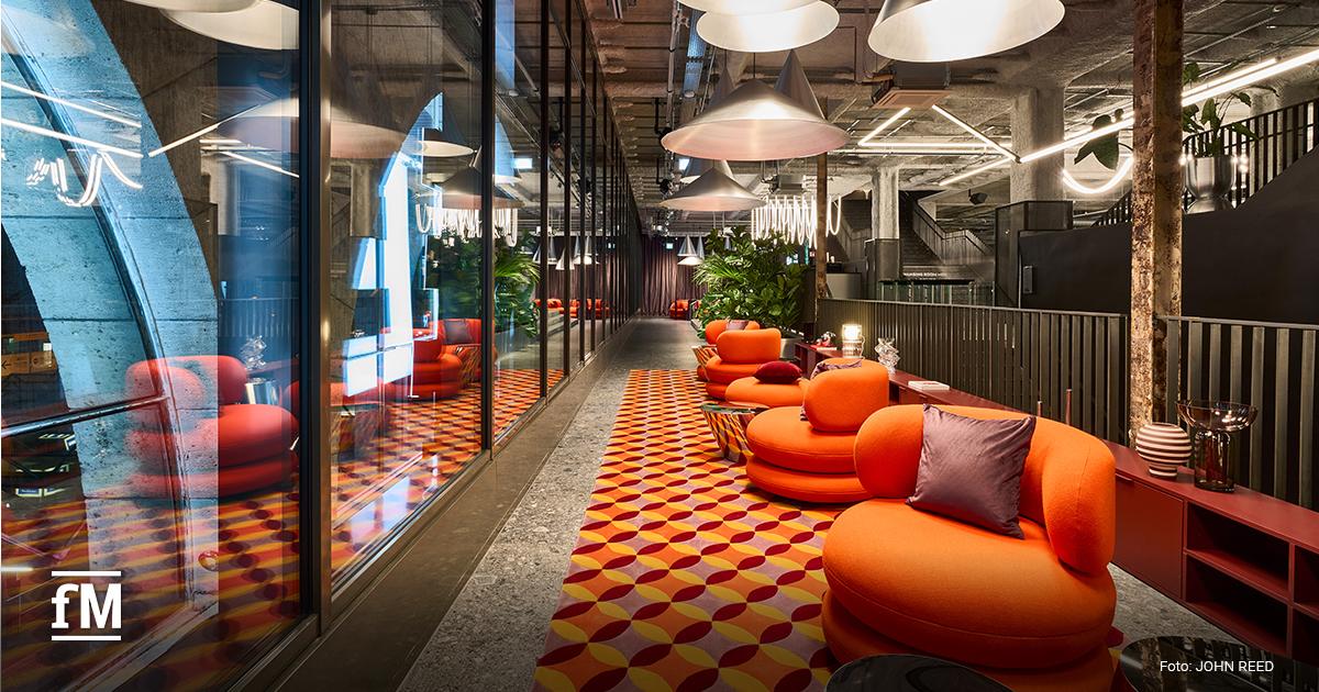 Stilvolle Fitnessstudio-Lounge mit orangefarbenen Rundsofas, geometrisch gemustertem Teppich, großen Glaswänden und industriellen Pendelleuchten.