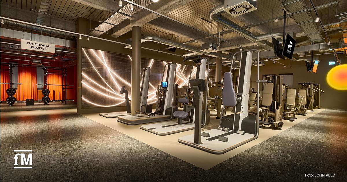 Großzügiger Fitnessbereich mit Kraft- und Cardiogeräten, breitem Mittelgang, industrieller Betondecke und geschwungenen LED-Lichtinstallationen.