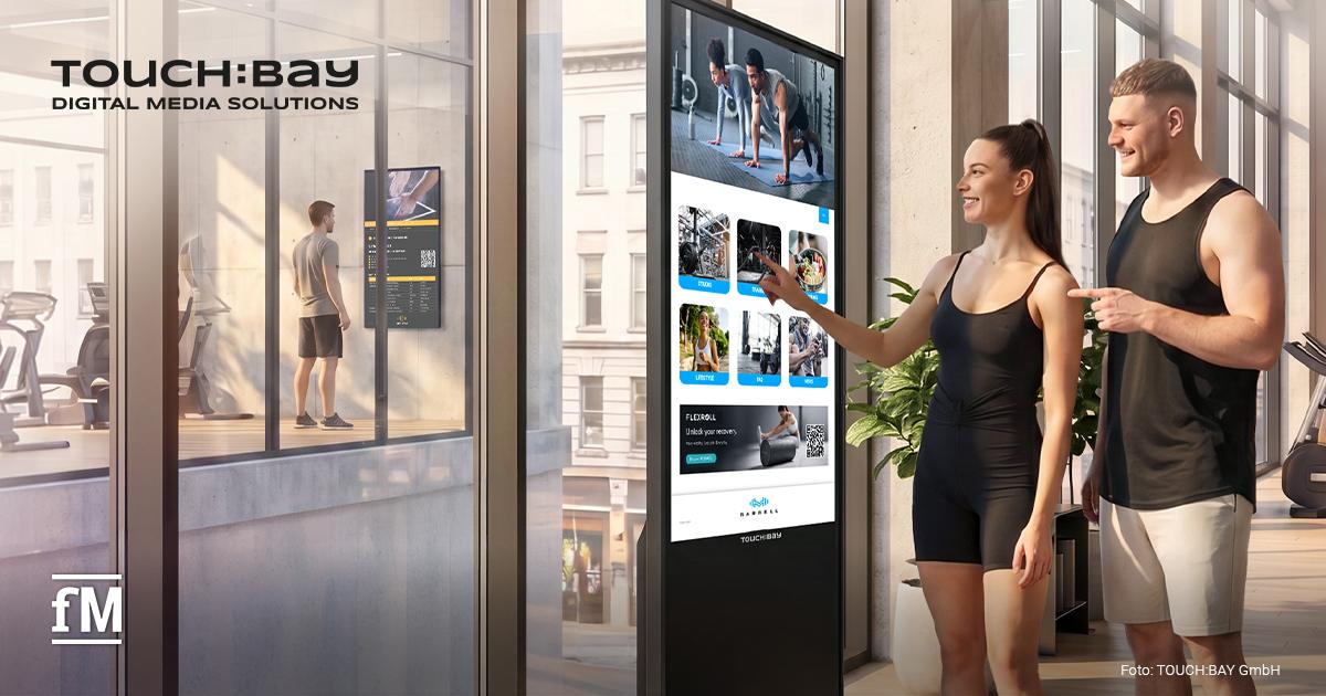 TOUCH:BAY Expansion Im Vordergrund bedienen eine Frau und ein Mann in Sportkleidung einen freistehenden digitalen TOUCH:BAY Bildschirm. Auf dem Display sind mehrere Kacheln mit Trainings- und Lifestyleinhalten sichtbar. Links hinter einer Glasfront steht eine Person vor einem weiteren Display. Oben links steht das Logo „TOUCH:BAY DIGITAL MEDIA SOLUTIONS“