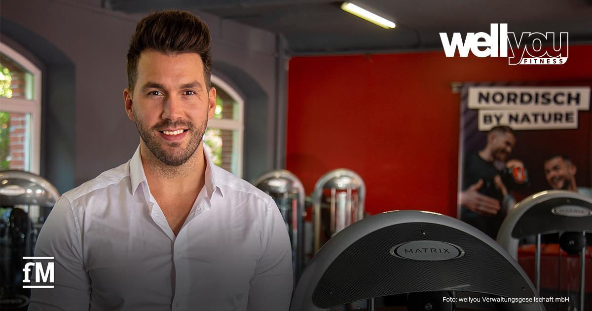 Ein Mann (Markus Sigl) in weißem Hemd steht in Fitnessstudio vor Trainingsgeräten; rechts oben Logo „wellyou Fitness“, links unten „fM“-Logo.