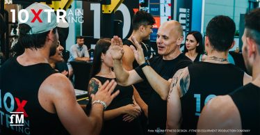 Gruppe von Menschen in Sportkleidung gibt sich im Fitnessstudio High-Fives. Oben links das Markenlogo von 10XTraining.
