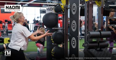 Frau wirft einen Medizinball in einem Fitnessstudio, weitere Trainierende und funktionelle Trainingsgeräte sind im Hintergrund zu sehen.