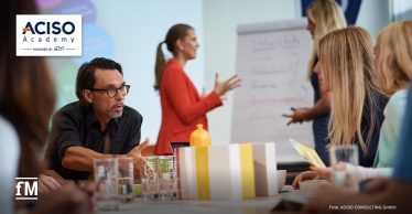 Menschen in einem Seminarraum bei einer Schulung der ACISO Academy; ein Mann spricht engagiert, während im Hintergrund eine Frau an einem Flipchart präsentiert.