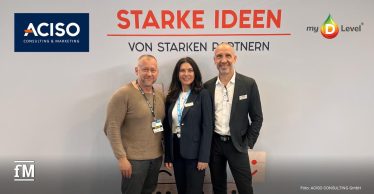 Drei Personen stehen frontal im Vordergrund vor einer hellen Messewand mit roter Überschrift „STARKE IDEEN“ und schwarzem Text „VON STARKEN PARTNERN“; die Personen tragen Messeausweise, im Hintergrund befinden sich Hallendecke, Beleuchtung und Messestanddetails sowie die Firmen-Logos von ACISO und my-D-Level®