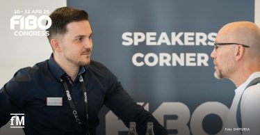 Zwei Männer im Gespräch beim FIBO Congress 2025. Der Mann links – Thorsten Clemann, Management-Dozent an der DHfPG/BSA-Akademie – trägt ein dunkles Hemd mit einem FIBO-Lanyard und hat eine Namensplakette an der Brust. Der Mann rechts, mit Brille und weißem Oberteil, hört aufmerksam zu. Im Hintergrund ist eine dunkelblaue Wand mit der Aufschrift 'SPEAKERS CORNER' und dem FIBO Congress Logo zu sehen. Das fM-Logo befindet sich unten links im Bild.