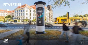 Freistehende digitale Werbesäule im Vordergrund auf einem Platz am Straßenrand. Links oben im Bild steht das Logo „FITVERTISE“, links unten das Logo „fM“. Im Hintergrund befinden sich historische Gebäude, eine gelbe Straßenbahn und eine Haltestelle. Unscharfe Passantinnen und Passanten bewegen sich links und rechts vor der Werbesäule.