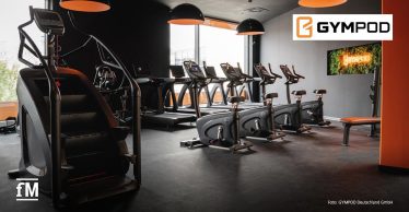 Innenansicht eines modernen Fitnessstudios mit Laufbändern, Fahrradergometern, Treppensteiger und Sitzbank; an der Wand ein Monitor mit GYMPOD-Logo.