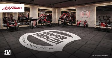 Moderner Trainingsraum mit zahlreichen roten und schwarzen Hammer Strength Geräten. In der Mitte des Raumes ist ein großer, runder Schriftzug 'Hammer Strength Training Center' auf dem Boden zu sehen.