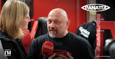 Interview-Szene auf einer Fitnessmesse: Eine Reporterin von fitness MANAGEMENT mit Mikrofon im Gespräch mit einem Mann im schwarzen Panatta-T-Shirt, im Hintergrund rote Fitnessgeräte.