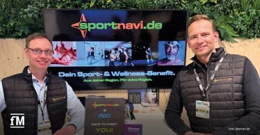 Reinhard Hunke und Hannes Hiemesch in schwarzen Sportnavi.de-Westen stehen lächelnd am FIBO-Messestand mit Bildschirm im Hintergrund, auf dem das Logo von sportnavi.de und der Schriftzug ‚Dein Sport- & Wellness-Benefit‘ zu sehen sind.
