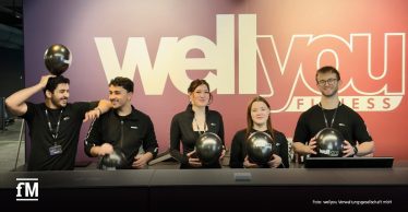 Fünf Mitarbeitende von wellyou stehen hinter einem Empfangstresen vor einer großen „wellyou Fitness“-Wand und halten schwarze Luftballons mit „wellyou“-Logo in den Händen. Links und rechts tragen die Personen schwarze Arbeitskleidung mit Namensbändern.