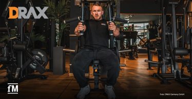 Im Vordergrund sitzt ein Mann an einem schwarzen Kraftgerät in einem Fitnessstudio und hält zwei Griffe auf Brusthöhe. Links oben steht das orange-graue Logo „DRAX“, links unten das weiße Logo „fM“, rechts unten der Text „Foto: DRAX Germany GmbH“.