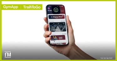 Zentrale Vordergrundansicht eines Smartphones in einer Hand, Display zeigt die App-Oberfläche mit Button „Eingang öffnen“, Bereich „Dein Studio“ mit Bild eines Fitnessraums, Menüleiste unten, oben links Schriftzug „GymApp | TrainToGo“, unten rechts Text „Foto: Gym App GmbH“.