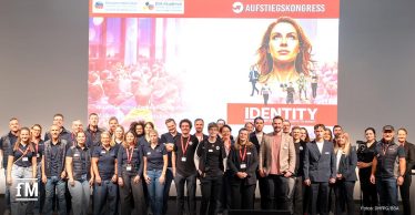 Gruppenfoto mit rund 30 Personen vor der Bühne des Aufstiegskongresses mit großem Veranstaltungsbanner im Hintergrund.
